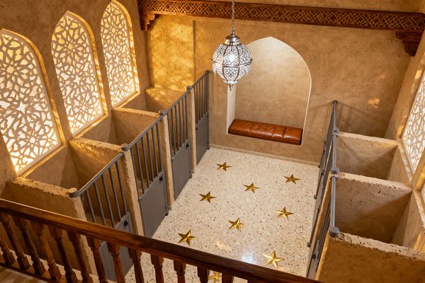 An elevated view of a Moroccan-inspired home gym shows sand-colored plaster walls with backlit arched jali screens and a large filigree metal pendant. A pale terrazzo floor is inlaid with brass stars, and narrow workout bays are divided by rough plaster half walls and gray steel railings with paneled doors. A built-in arched alcove holds a caramel leather bench, while carved wood corbels and a lattice cornice add detail.