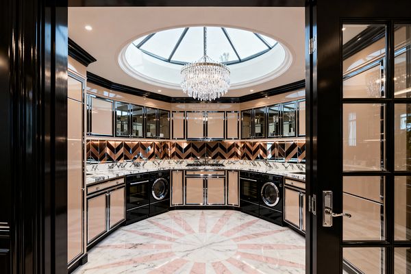 Luxurious circular laundry room viewed through glossy black French doors, centered beneath a large domed skylight with a crystal chandelier. Curved walls hold mirrored upper cabinets and champagne-lacquer lower cabinets with chrome trim, a rose-gold and black chevron mirrored backsplash, white marble countertops, integrated black front-loading appliances, and a white marble floor with a blush-pink sunburst inlay, all highlighted by warm under-cabinet lighting and black crown molding.