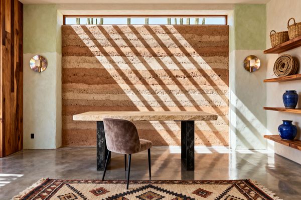 A Southwestern-inspired home office features a thick stone desk on black metal supports placed before a striped rammed-earth wall. Polished concrete floors, a geometric kilim rug, limewashed sage and cream plaster, solid wood shelves with woven baskets and cobalt-blue pottery, circular metal sconces, and a narrow clerestory window casting diagonal sunlight complete the warm, minimal desert look.