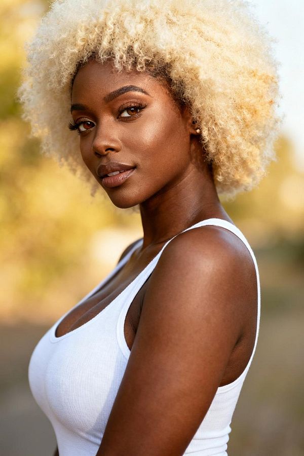 A portrait of a woman with curly blonde hair, wearing a white tank top, set against a blurred natural background.
