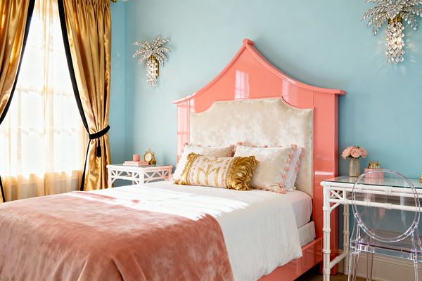 A kids’ bedroom in Hollywood Regency style features aqua walls and a glossy coral pagoda headboard with an ivory velvet upholstered panel. The bed has white bedding, a blush velvet throw, pom-pom pillows, and a gold-fringed bolster. Crystal palm-tree sconces flank the bed. Gold satin drapes with black trim filter daylight, while a white faux-bamboo desk and nightstand sit beside a clear acrylic chair and a vase of pink roses.