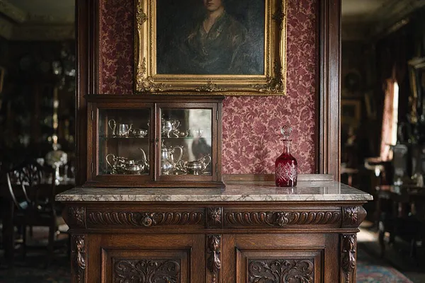 A Victorian-style interior vignette featuring a carved dark wood cabinet with a marble top, a glass-front display case of silver serving pieces, and a small red cut-glass decanter. Pink damask wallpaper and a gilt-framed portrait create a rich, formal atmosphere.
