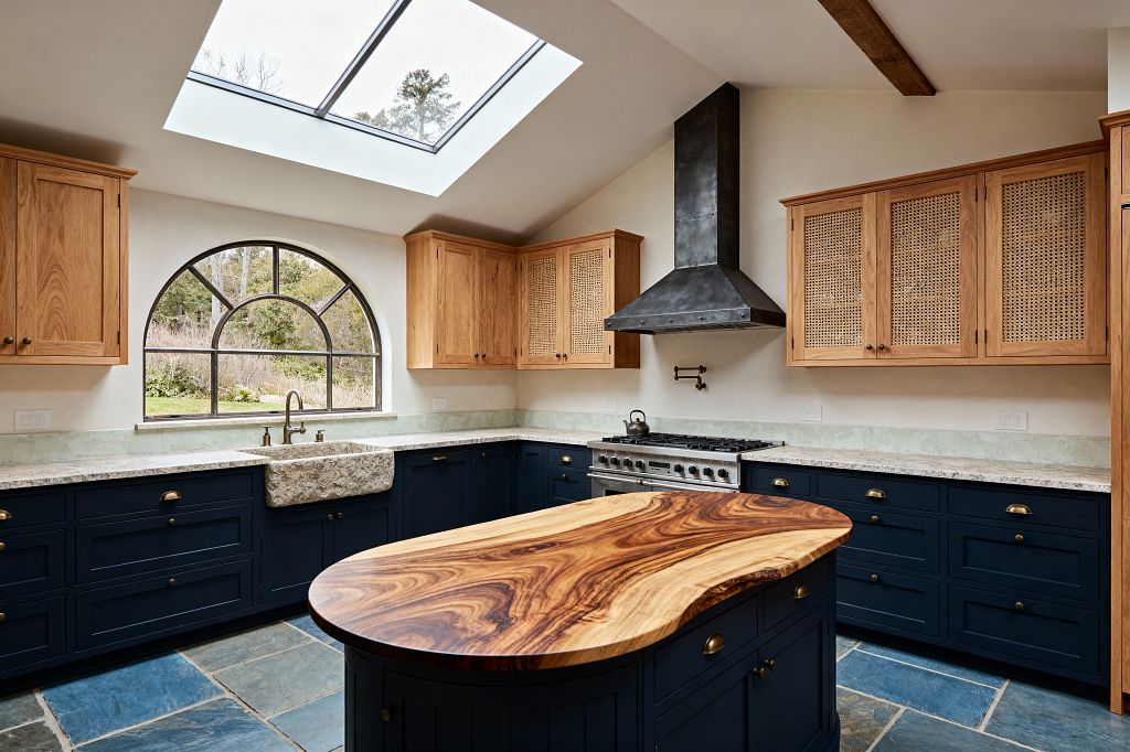 A sunlit transitional kitchen with deep navy Shaker base cabinets and brass pulls, topped with light stone countertops. Upper cabinets in natural oak feature cane panels. A rough-hewn stone farmhouse sink sits beneath an arched black-steel window, with a large skylight above. A blackened steel range hood, pro-style range, and brass pot filler line one wall. The island has a sculptural live-edge wood top, and the floor is blue-gray slate tile.