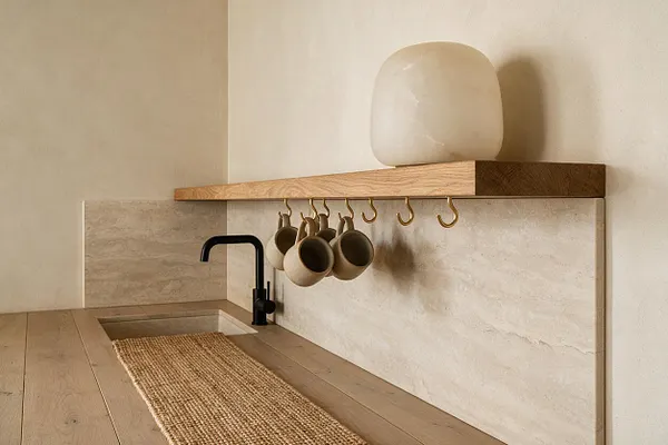 A serene Organic Modern kitchen corner with a matte black faucet, light oak countertop, and pale stone backsplash and sink surround. Open brass hooks hold small neutral-toned cups beneath a floating wood shelf topped with a sculptural cream vessel, while a woven runner adds natural texture.
