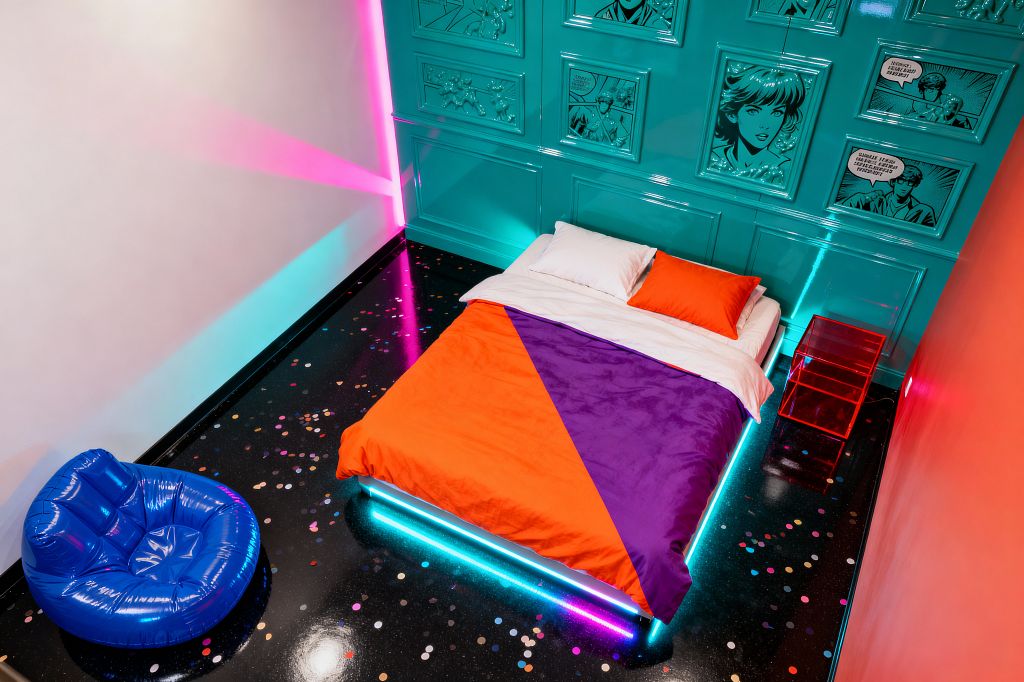 A Pop Art bedroom with a glossy teal paneled feature wall displays framed comic-style graphics above a low bed edged with cyan and magenta LED lights. The bed is dressed in orange and purple geometric bedding with white pillows, beside a translucent red acrylic nightstand; a cobalt blue vinyl lounge chair and a polished black epoxy floor scattered with confetti-like chips complete the space, while neon light washes color the white and coral walls.
