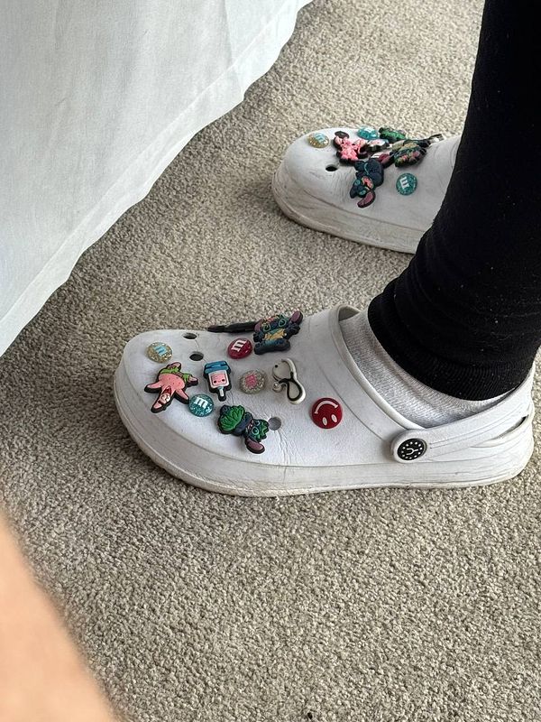 A close-up of a decorated white Crocs shoe resting on a carpeted surface.