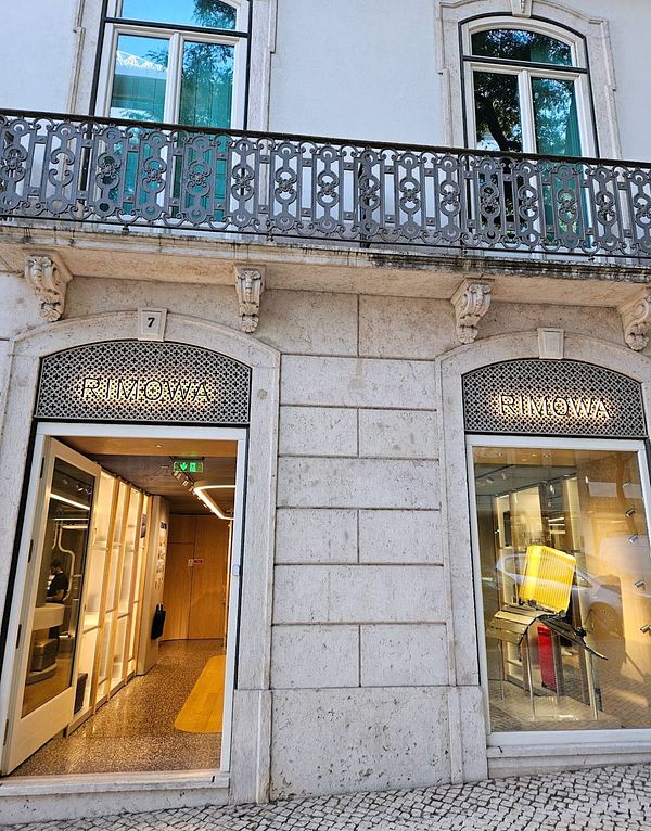 The image depicts the exterior of a Rimowa store in Lisbon, showcasing its entrance and window displays.