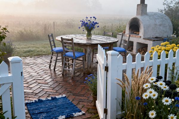 A misty farmhouse patio features a round, weathered wood table surrounded by ladder-back chairs with blue gingham cushions, set on dew-damp herringbone brick. A white picket gate opens to a domed stucco wood-fired oven with stacked logs below; a fringed blue mat and cottage plantings of daisies, cornflowers, yellow blooms, and wheat frame the scene, all bathed in soft sunrise light with a meadow beyond.