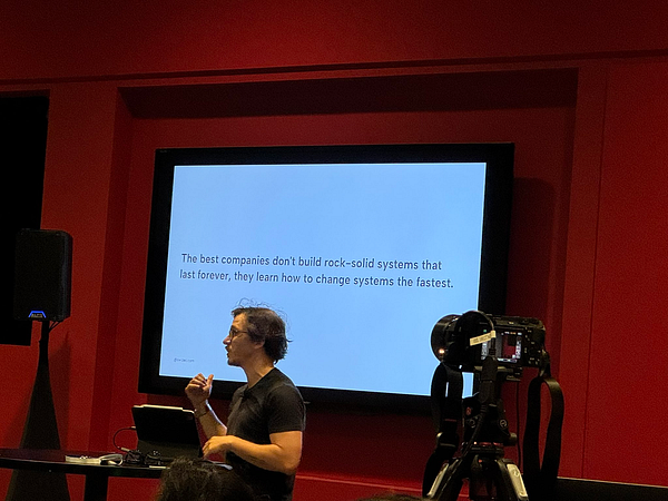 A speaker presents a talk in front of an audience, with a quote displayed on a screen.