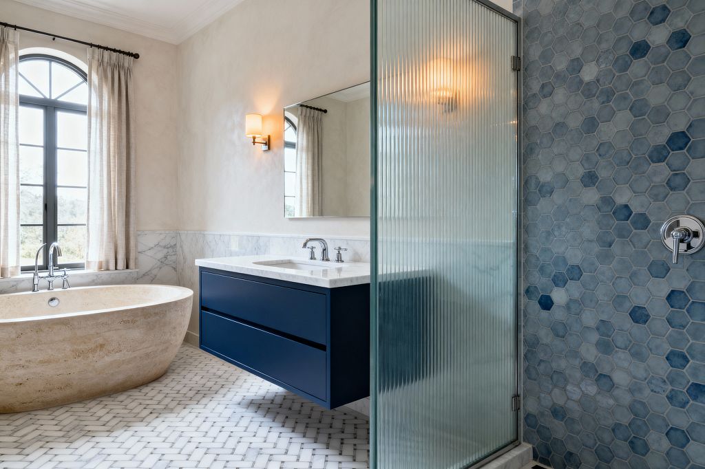 A transitional bathroom with a freestanding oval travertine tub set beneath an arched black-framed window with beige curtains. A navy floating vanity with a white marble countertop and polished-nickel faucet sits below a frameless mirror and a warm wall sconce. The floor is white marble basketweave mosaic, walls feature Carrara marble wainscot with plaster above, and a shower with ribbed glass reveals blue hexagonal tiles and a chrome control.