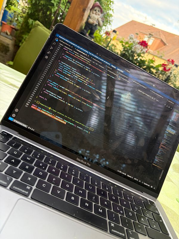 A person coding on a laptop outdoors under sunny weather, surrounded by greenery and a relaxing environment.