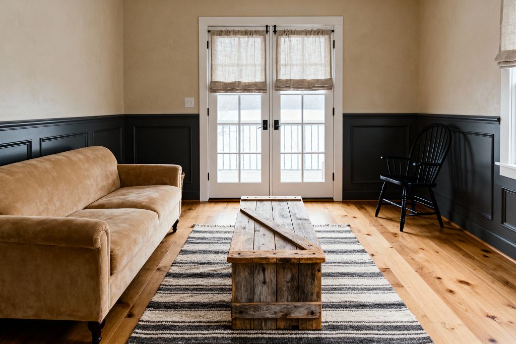 A farmhouse living room shows a camel-colored sofa on the left, a weathered reclaimed-wood trunk used as a coffee table centered on a black-and-cream striped rug, and a black Windsor chair in the right corner. Honey-toned wide plank floors lead to white-trim French doors with black hardware and light linen Roman shades. The walls are beige above deep charcoal paneled wainscoting, and daylight softly fills the uncluttered space.