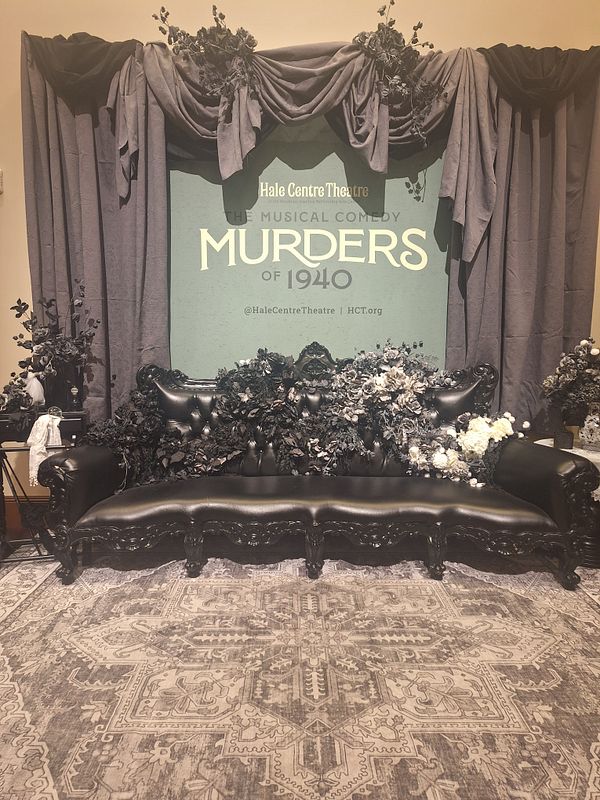 A decorative theatre backdrop featuring a black couch and floral arrangements for the play 'Murders of 1940'.