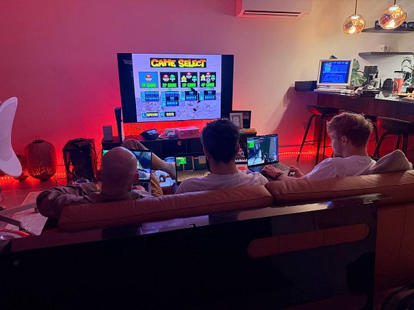 Three people sitting on a sofa playing video games with a TV screen and their personal laptops in a room lit with red LED lights.