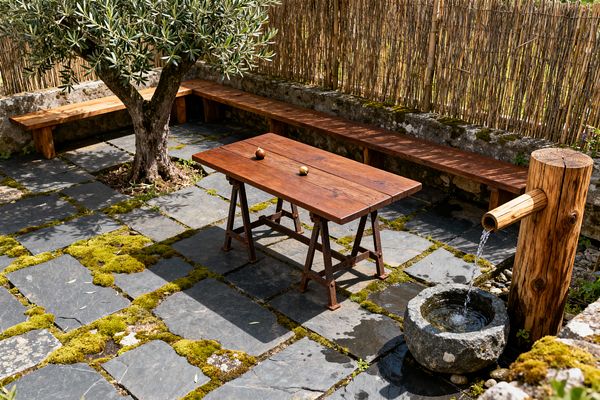 Rustic patio with charcoal slate pavers threaded with moss, centered by a narrow plank table on rusted metal trestles. A long reclaimed-wood bench lines a mossy stone wall and bamboo reed screen, while a log post feeds a wooden spout into a round carved stone basin; an olive tree casts dappled shade.