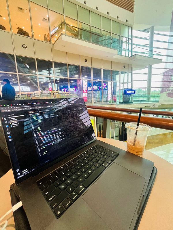 A laptop displaying code is set on a table in a mall with a view of the interior.