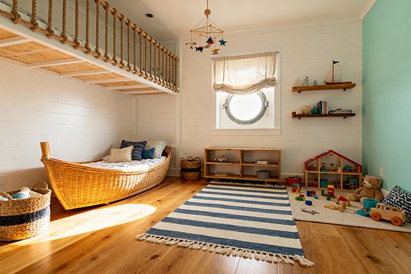 Children's room in coastal style featuring a boat-shaped wicker bed under a loft with thick rope railing and light wood slats. White shiplap walls, a seafoam accent wall, and a round porthole window with a linen Roman shade frame a space with honey-toned wood floors, a blue-and-cream striped rug, low cubby storage, floating shelves with a model sailboat, and a play corner with blocks, a dollhouse, and plush toys, all lit by warm natural sunlight.