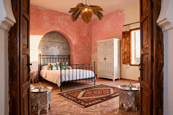 A Moroccan-style bedroom features blush pink plaster walls and a herringbone terracotta floor, centered on an iron bed set inside an arched niche clad in blue-and-white zellige tiles. Sunlight streams through wooden shutters onto a carved white armoire, white bedding with cactus-print pillows, a red geometric kilim rug, and two silver hammered tea tables, while a palm-leaf pendant hangs from the ceiling.