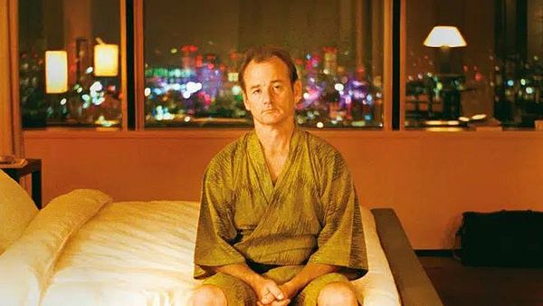 A man sits on a bed in a hotel room, looking contemplative against a vibrant city backdrop at night.