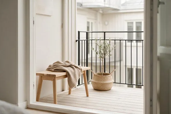 A serene Scandinavian-style balcony with pale wood decking, a simple light wood bench draped with a beige throw, and a woven basket planter holding a small olive tree. Black metal railings contrast with the soft white exterior walls and bright, airy surroundings.