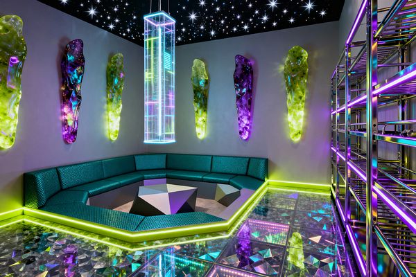 Media room with a sunken hexagonal sofa in teal textured upholstery around a faceted low table. Gray walls display sculpted resin artworks that glow in lime green and purple. Above, a black ceiling dotted with fiber‑optic stars frames a tall prismatic acrylic pendant. The floor is glass with holographic facets and neon edge lighting, reflecting the colors. At right, iridescent stainless‑steel shelving is lined with RGB strips.