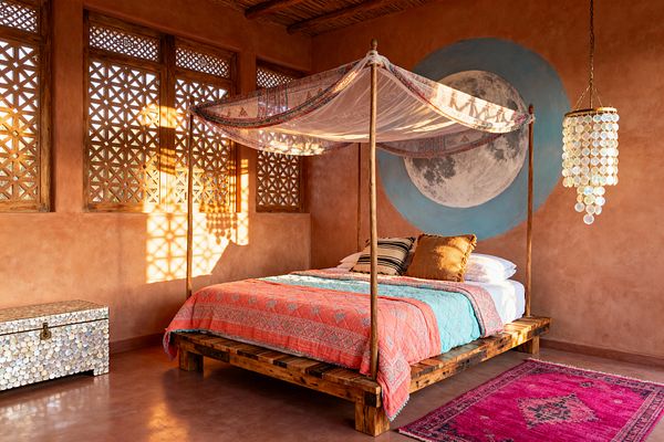 A bohemian bedroom with terracotta plaster walls features a low reclaimed-wood canopy bed draped in sheer patterned fabric and layered coral and turquoise quilts. A large moon mural with a pale blue halo anchors the bed. Sunlight pours through wooden lattice windows, casting geometric shadows on the clay floor and bright fuchsia kilim rug. A tiered capiz shell pendant hangs beside the bed, with a matching mother-of-pearl trunk positioned under the windows.