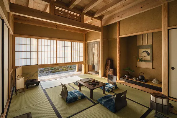 A serene Japanese Zen sunroom with tatami mat flooring, low wooden tea table, and blue floor cushions arranged for sitting on the floor. Shoji sliding screens fill the back wall with soft daylight, while natural wood beams, an alcove with calligraphy and a teapot display, and earthy walls create a calm, minimalist atmosphere.