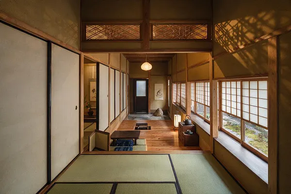 A tranquil Japanese Zen entryway with tatami flooring in the foreground, wood-framed shoji screens, and a long corridor of warm natural timber. Soft lantern-style lighting, a low seating area, and garden views through paper-paneled windows create a calm, minimalist atmosphere.