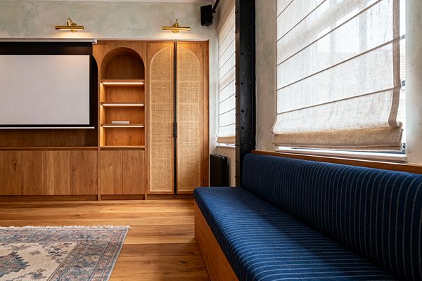 A modern farmhouse media room with a built-in oak wall: a pull-down projection screen at left, open arched shelving with integrated LED strips, and cane-front arched cabinet doors beneath brass picture lights. To the right, large windows with beige linen Roman shades sit above a long navy pinstripe upholstered bench on an oak base, with wide-plank wood flooring, a vintage fringed rug, and a matte black steel column and radiator.