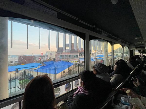 Passengers are seated inside a train, looking out at a seaside view with blue rooftops and distant buildings.