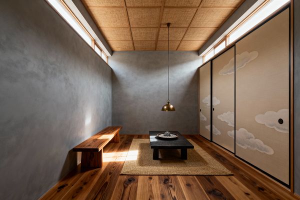 A minimalist Japanese Zen living room with smooth gray plaster walls and high clerestory windows admitting soft daylight. A low matte-black table rests on a woven jute rug beneath a small brass pendant, with a long solid-wood bench along the left wall. Warm knotty wood flooring and a wood-grid ceiling filled with woven natural-fiber panels add texture. On the right, beige fusuma sliding doors edged in black display white cloud motifs, reinforcing a tranquil, uncluttered atmosphere.