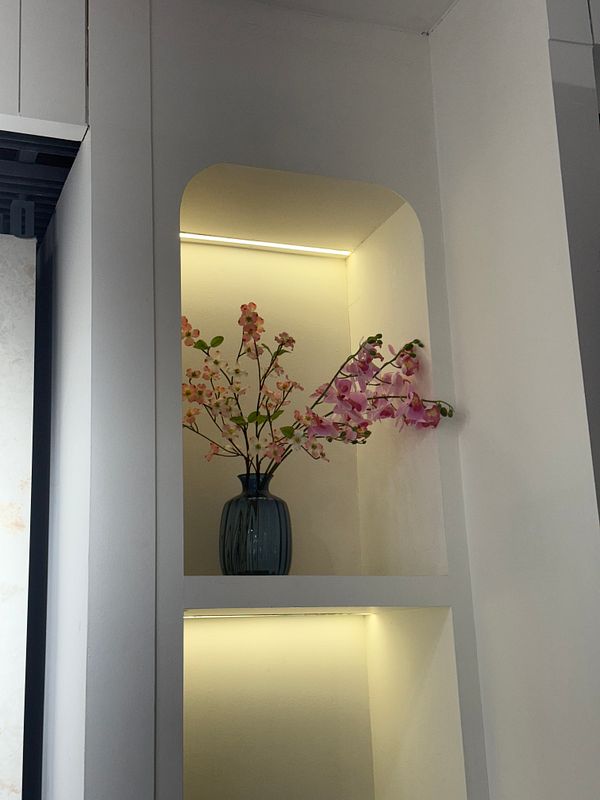 A decorative shelf features a vase with artificial flowers, illuminated by soft lighting.