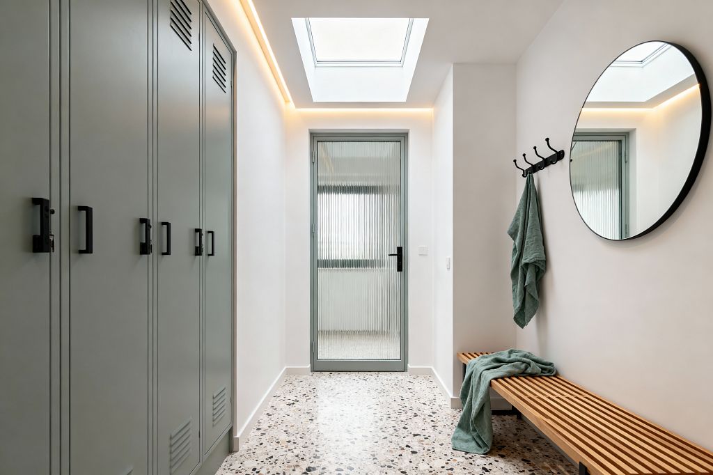 A bright contemporary mudroom features sage-gray built-in lockers with black handles on the left and a slatted oak bench with black legs on the right. Speckled terrazzo flooring leads to a ribbed-glass door under a skylight with warm cove lighting; a large round black-framed mirror and a row of black hooks hold muted green towels against white walls.