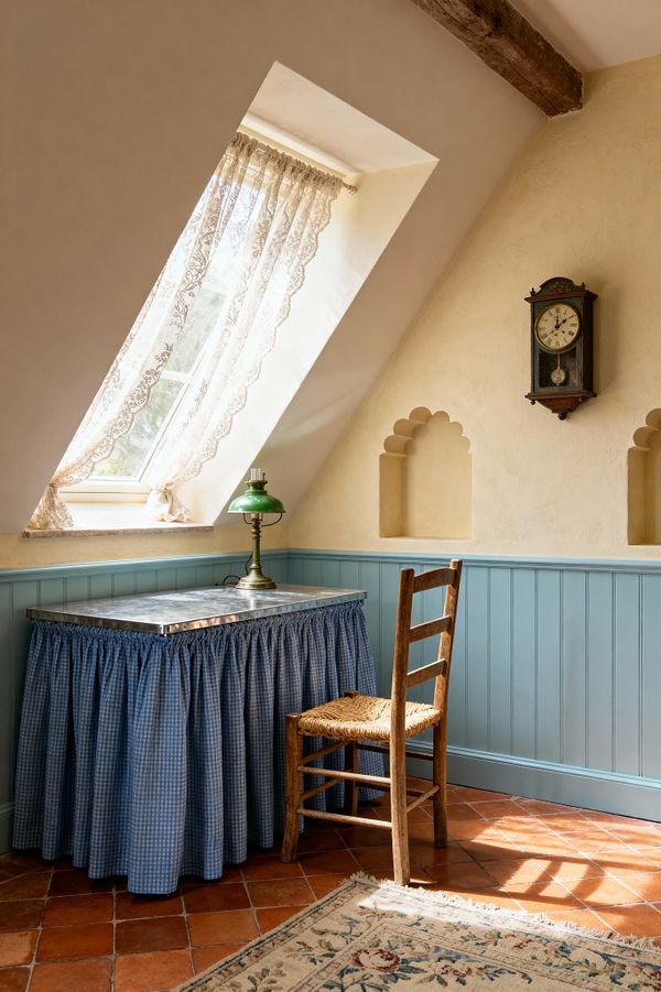 A small attic home office features a sloped dormer window with lace curtains, cream plaster walls, and powder-blue beadboard wainscoting. A skirted desk in blue gingham with a zinc top holds a brass lamp with a green glass shade. A rustic ladder-back chair with a woven rush seat sits on terracotta tile flooring beside a floral rug. Carved wall niches, a vintage pendulum clock, and an exposed timber beam complete the cozy, sunlit space.