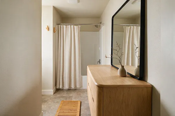 A serene master bathroom with a light oak vanity, a large black-framed mirror, and a neutral palette of cream and beige. A softly textured shower curtain, stone-look floor tiles, and a small wooden bath mat complete the calm Japandi-inspired space.
