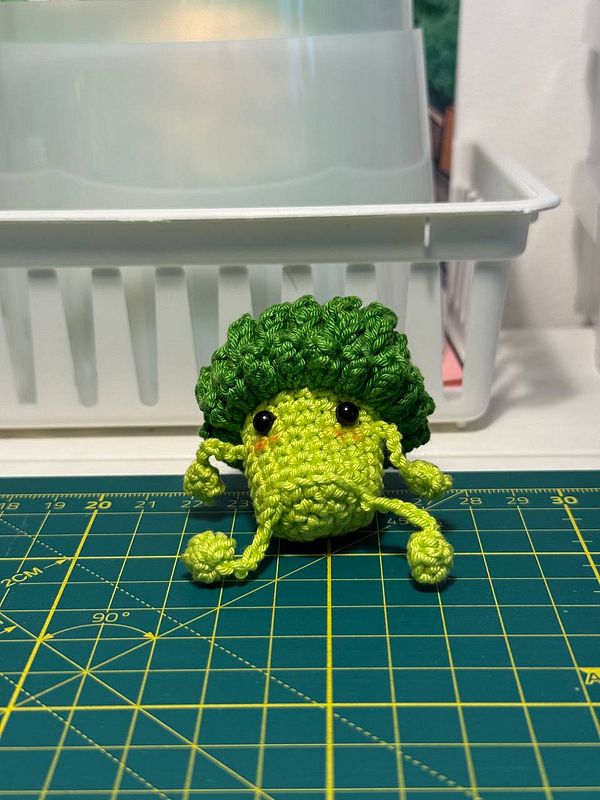 A crocheted green critter resembling a broccoli with a cheerful expression is displayed on a cutting mat.