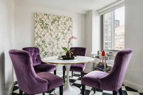 A bright game room features four purple velvet dining chairs arranged around a round white pedestal table with a gold rim. The room has black-and-white checkered flooring, a floral lattice wall panel, and a window with a small acrylic and chrome bar cart beside it.