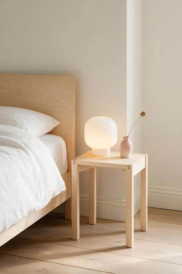 A serene Scandinavian bedroom vignette with a light wood bed frame, white textured bedding, and a small birch side table. The table holds a softly glowing frosted glass lamp and a pale pink vase with a single dried stem beside a cream wall and pale wood floor.
