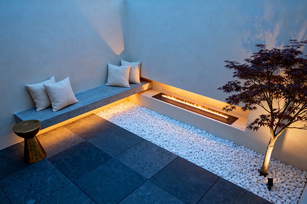 A compact walled patio at dusk features a floating concrete bench with light linen pillows along a smooth plaster corner. Warm LED strips glow beneath the seat and within a narrow recessed linear fire set into the low wall. Large charcoal stone pavers edge a bed of crisp white river pebbles, where a small Japanese maple is up-lit from below. A sculptural brass side table sits beside the bench, emphasizing the minimalist palette and clean geometry.