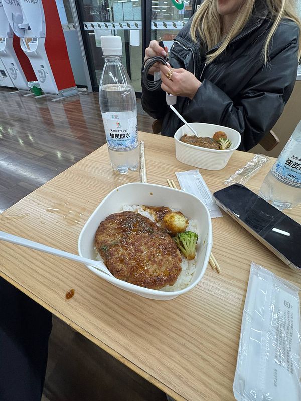 A meal from 7/11 featuring a rice bowl with a meat patty and vegetables, accompanied by a bottle of water.