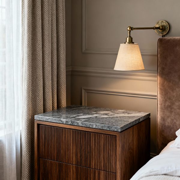 A close-up of a transitional bedroom corner shows a walnut nightstand with vertically reeded drawers and a gray marble top beside a tall taupe upholstered headboard. Above, an antique brass wall sconce with a tapered linen shade glows warmly. Greige paneled walls provide classic detailing, while layered window coverings—a sheer white curtain and a beige herringbone drape—filter daylight. The palette is neutral and soft, blending traditional and contemporary elements.