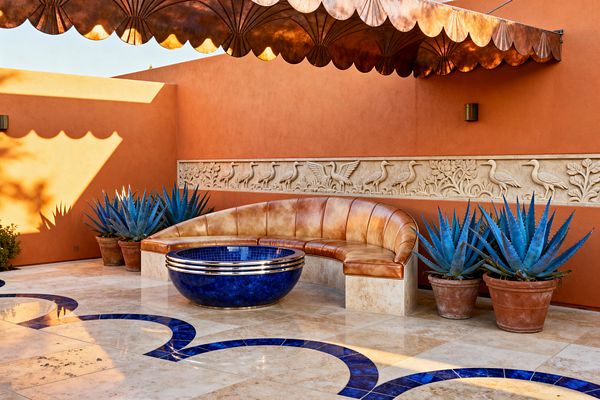 A terracotta-walled patio features a scalloped copper awning casting fan-shaped shadows over a carved stone frieze of birds. At center, a curved caramel leather banquette on a travertine plinth faces a low circular bowl clad in deep cobalt mosaic with polished metal bands, set on a cream stone floor with matching cobalt inlay; potted blue agaves and small brass sconces complete the Art Deco scene.