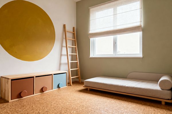 A minimalist children’s room features cork flooring, a light pine storage bench with three drawers in terracotta, rust, and dusty blue, each with oversized round pulls. A low beige daybed with a bolster sits beneath a window dressed with a linen Roman shade; a wooden ladder leans against a sage green wall, and a large mustard circle is painted on the adjacent white wall for a calm, sunlit feel.