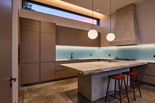 A contemporary kitchen features matte taupe, handleless cabinets in a U shape around a large island topped with a rough-cut stone slab on a concrete base. A pale aqua back-painted glass backsplash runs the perimeter with undercabinet LEDs; a sculptural concrete range hood anchors a gas hob, a flush black cooktop sits in the island, and two opal globe pendants, a clerestory window, and cove lighting illuminate polished concrete flooring and two reddish wood stools.