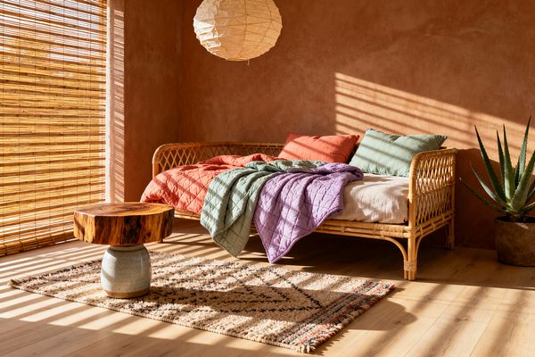 Sunlit living room with terracotta plaster walls and bamboo blinds casting striped shadows over pale wood floors. A rattan daybed holds a linen cushion, coral and sage pillows, and quilted throws in sage and lavender. A round paper lantern hangs overhead, a low table with a live-edge wood top sits on a tufted Moroccan-style rug, and an aloe plant in a rustic planter completes the natural, relaxed Bohemian look.