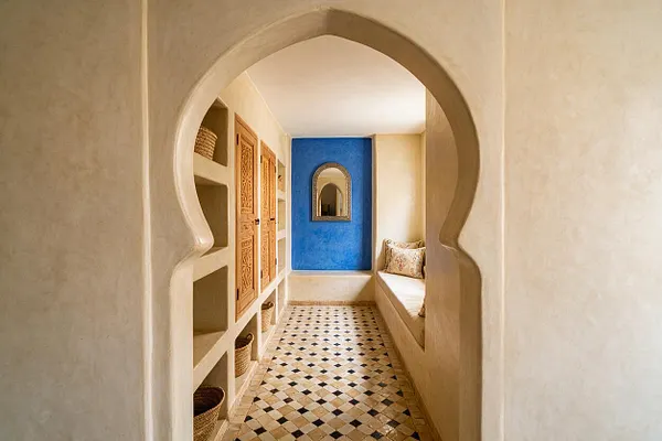A narrow Moroccan-style closet with a sculpted horseshoe arch framing the entrance, built-in niches and carved wooden cabinet doors on the left, and a cushioned bench with patterned pillows on the right. The space ends with a vivid blue accent wall, an ornate mirror, woven baskets, and a geometric tiled floor in warm neutral tones.