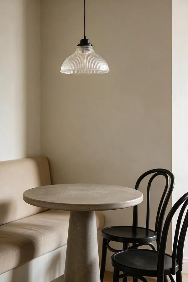 A small modern dining nook with a round pedestal table in a light stone finish, paired with a beige upholstered bench and two black bentwood-style chairs. A clear ribbed glass pendant hangs overhead, and the space is framed by soft neutral walls and a calm, minimalist palette.
