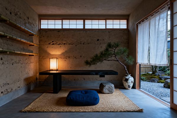 A minimalist Japanese Zen home office with earthen plaster walls and a low, ebonized wood desk illuminated by a warm paper lantern. A woven jute mat and deep indigo floor cushion rest on a polished concrete floor; bamboo shelves run along the left wall, while a pine branch in a crackle-glazed ceramic vase and a natural stone sit to the right, with shoji screens and sheer linen curtains opening to a small gravel and moss garden.