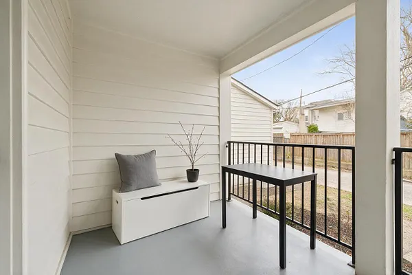 A small minimalist front porch with white horizontal siding, a light gray floor, and a simple black metal railing. A white storage bench with a gray cushion and small potted twig arrangement sits beside a black table, creating a clean and calm outdoor space.