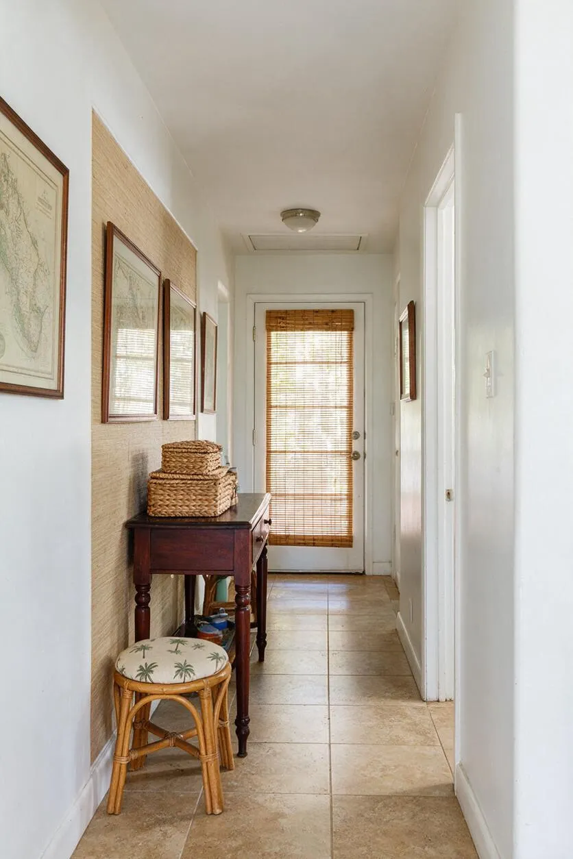 A narrow entryway with white walls and beige tile flooring features a dark wood console table on the left, topped with woven baskets. Framed maps and a textured woven wall panel add visual interest, while a bamboo stool with palm-print cushion sits in front. At the end of the hall, a door with woven bamboo blinds filters warm natural light.